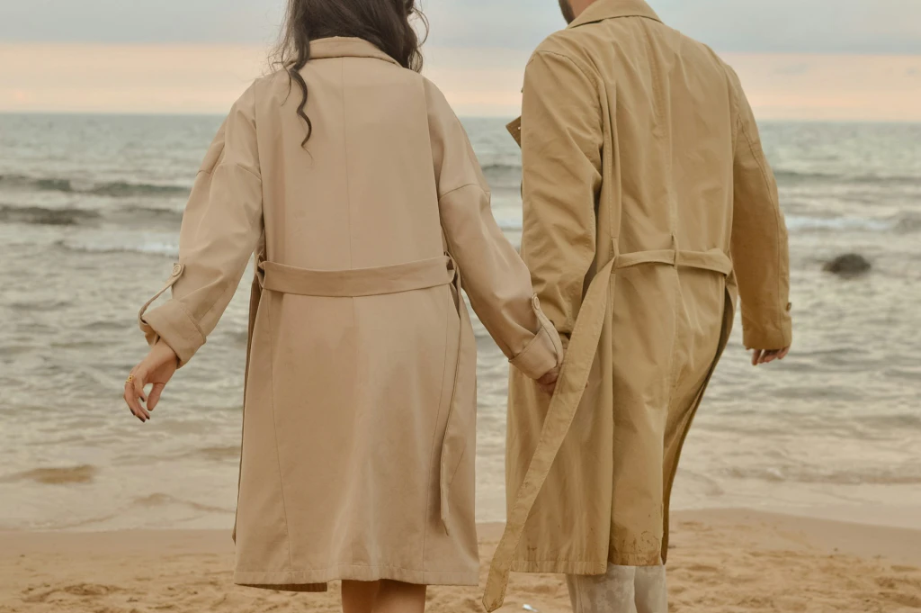 가을 아우터 트렌치코트를 입고 해변을 손잡고 걷는 두 사람의 뒷모습